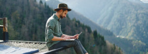 Man sieht einen Mann, der auf einem Vorsprung sitzt, auf ein Mobiles-Endgerät blickt und seine Beine in ein Tal baumeln lässt. Im Hintergrund sieht man mächtige Kiefern bewachsene Berge.