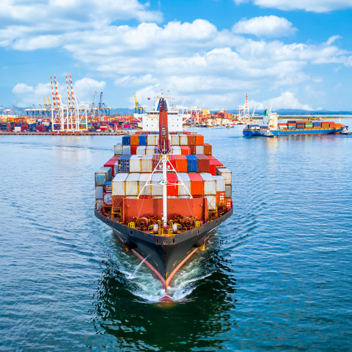 Containerfrachtschiff von vorne, im Hintergrund ein Hafen. In der Totalen Perspektive.